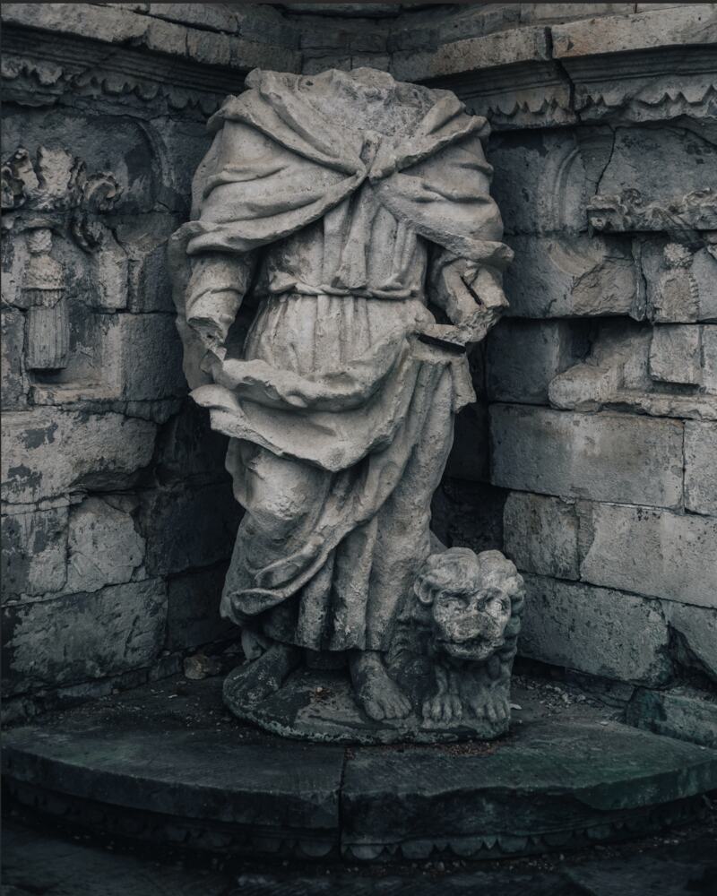 Source photograph: a weathered stone statue of a robed figure standing in a niche, with a carved lion at her feet.